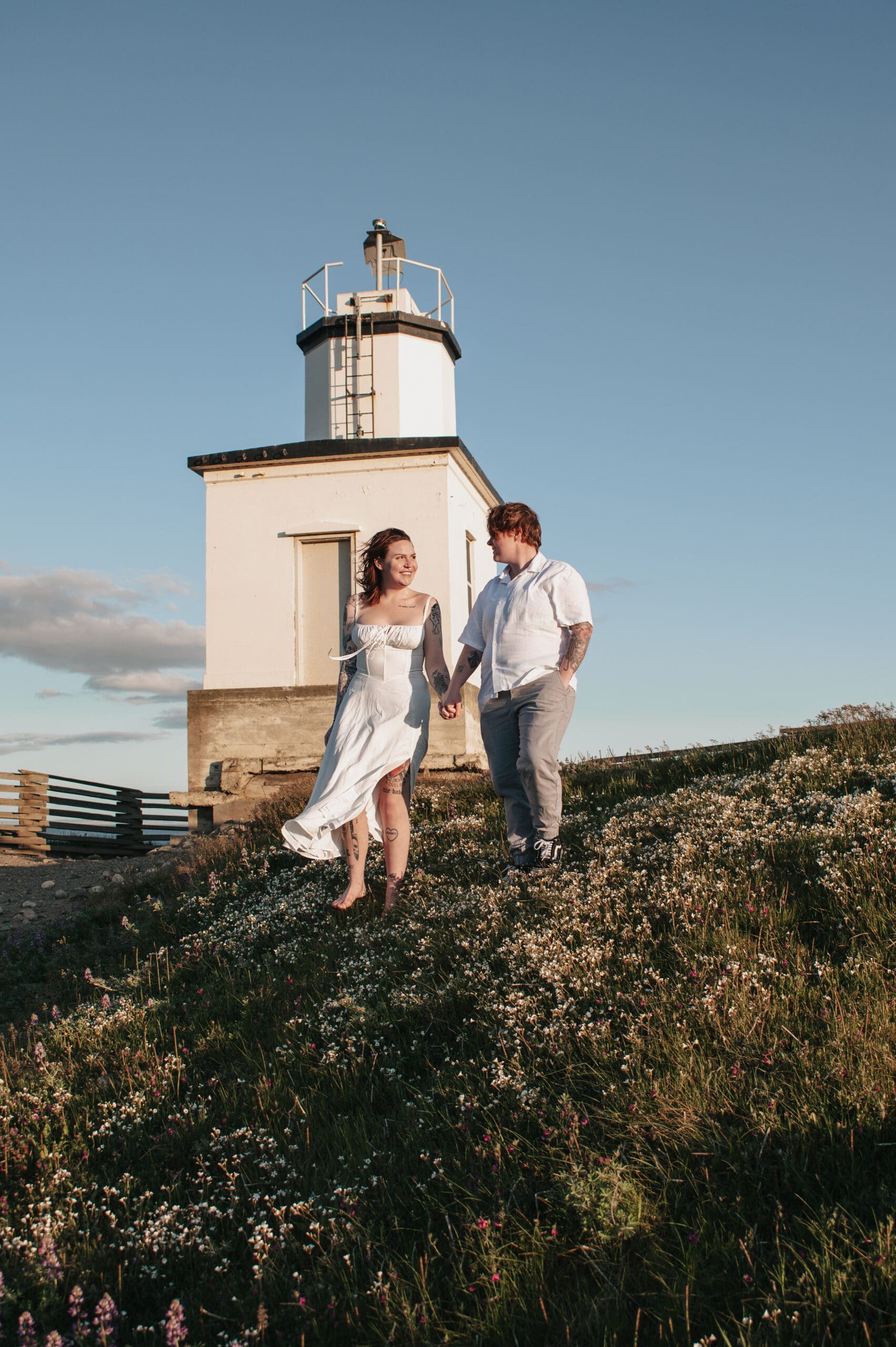 Engaged couple holding hands in front of Cattle Point Lighthouse during golden hour in the San Juan Islands surrounded by spring wildflowers.
