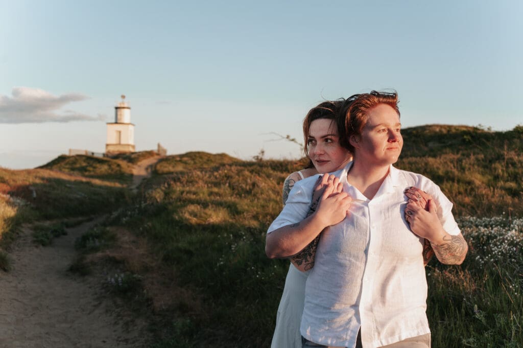 Golden light surrounds couple embracing with Cattle Point Lighthouse on the San Juan Islands in the background during destination engagement session