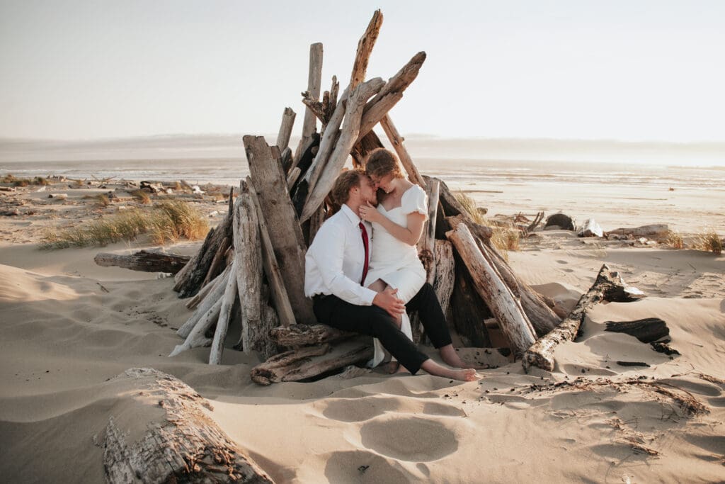 Engaged couple smiles while sitting in front of a driftwood teepee on the Oregon Coast
