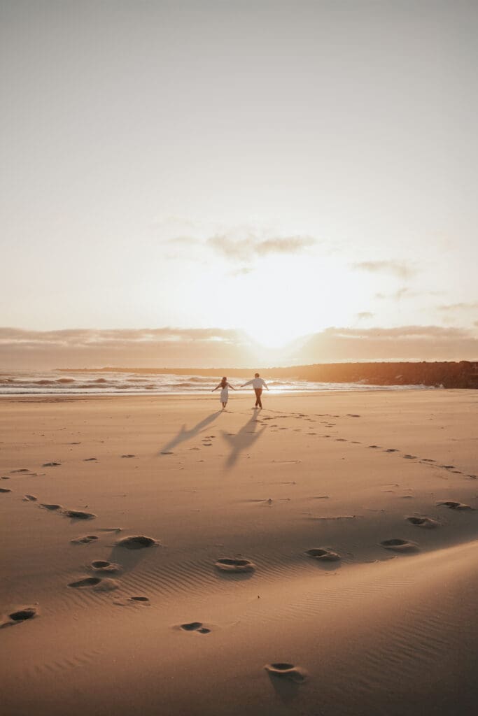 Couple runs hand-in-hand toward the shoreline after running in a heart shape during golden hour on the Oregon Coast
