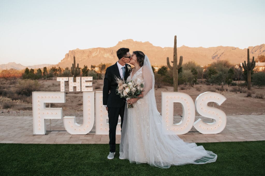 Bride and groom standing in front of Desert View wedding venue with custom marquee sign and the Superstition mountains behind them.