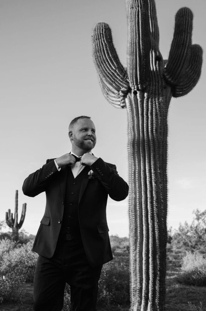 Black and white photo of groom walking and adjusting his bowtie with 2 big saguaro cactuses. 