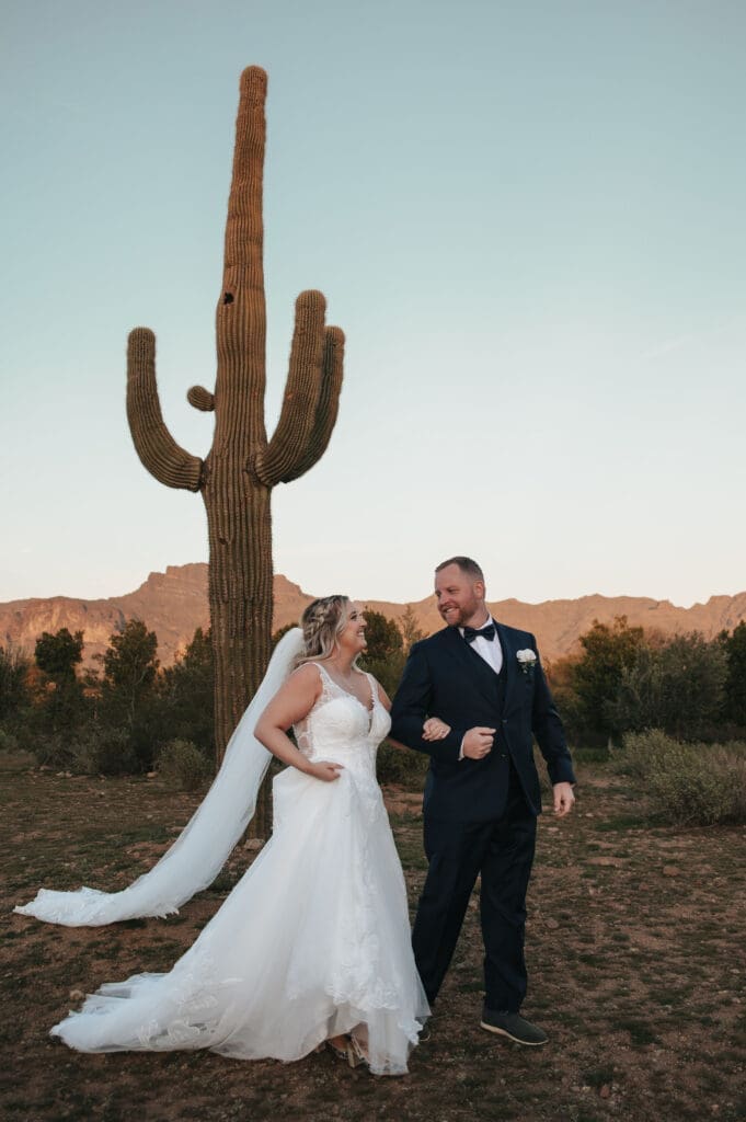Bride and Groom walk happily together in front of a Saguaro at the iconic Desert View Weddings and Events with views of the Superstition Mountains behind. 