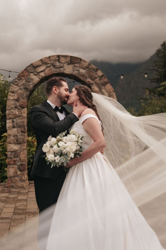 Bride and groom kiss in front of stone arch at Cape Horn Estate while bride's veil elegantly flows in the breeze.