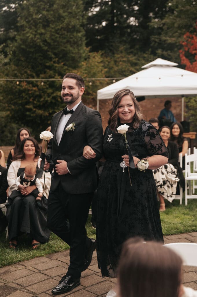 The Groom and his mother smile at guests while each holding a single white rose and walking down the aisle captured by Scottsdale Arizona Wedding Photographer.