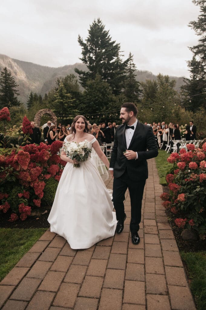 Bride and groom walking together smiling and holding hands while exiting their wedding ceremony through floral blooms at Cape Horn Estate.