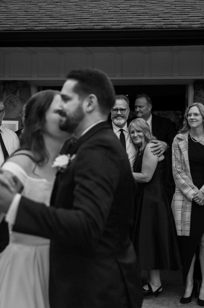 Black and white image of bride and groom dancing while parents watch emotionally in the background