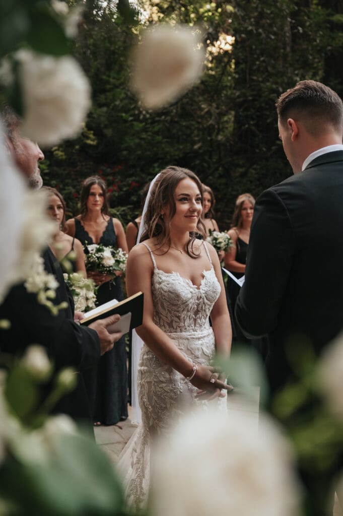 Bride smiling at groom during his vows to her during Nestldown wedding ceremony surrounded by flowers. 