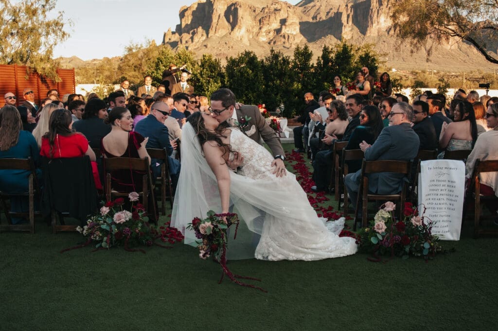 Couple embraces in a dip kiss while exiting outdoor wedding ceremony with mountain views at The Paseo Venue in Arizona in the background.