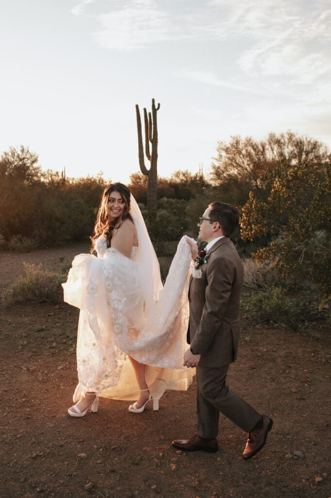 Groom helps carry train while bride and groom walking together near the Superstition Mountains at The Paseo Venue wedding at golden hour.