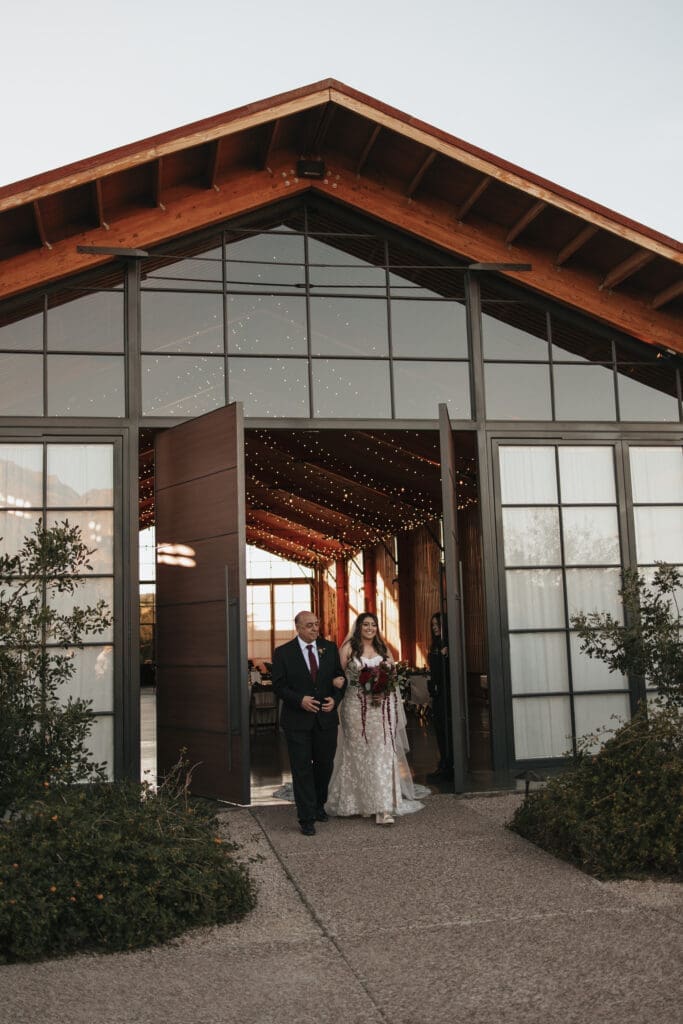 Bride walks out of building with her father during her wedding at the luxury, Award-Winning Paseo Venue in Apache Junction, AZ.